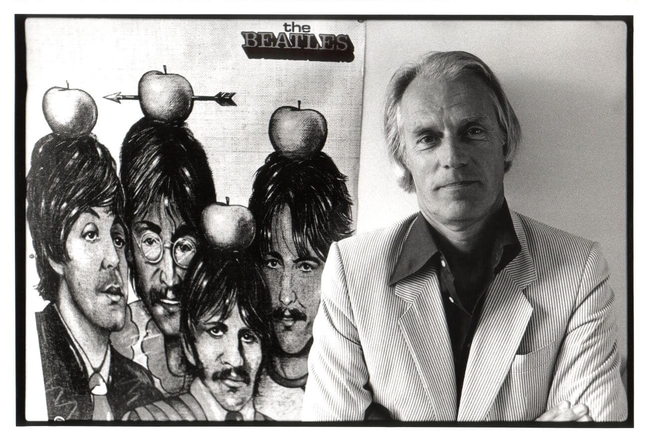 A black and white picture of George Martin posing with a drawing of The Beatles with apples on their heads.