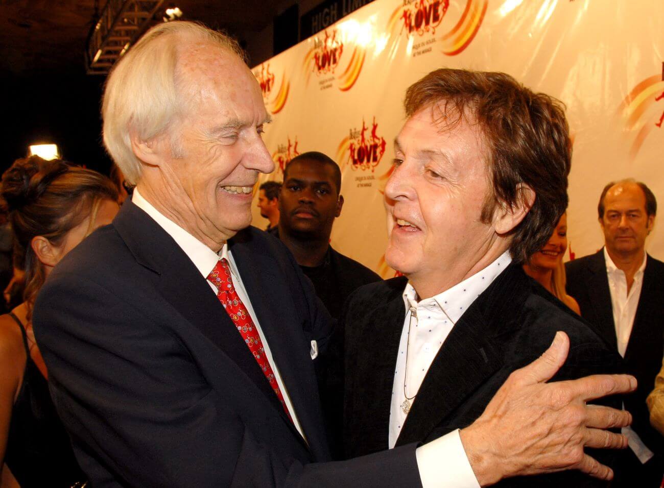 George Martin and Paul McCartney embrace on a red carpet.