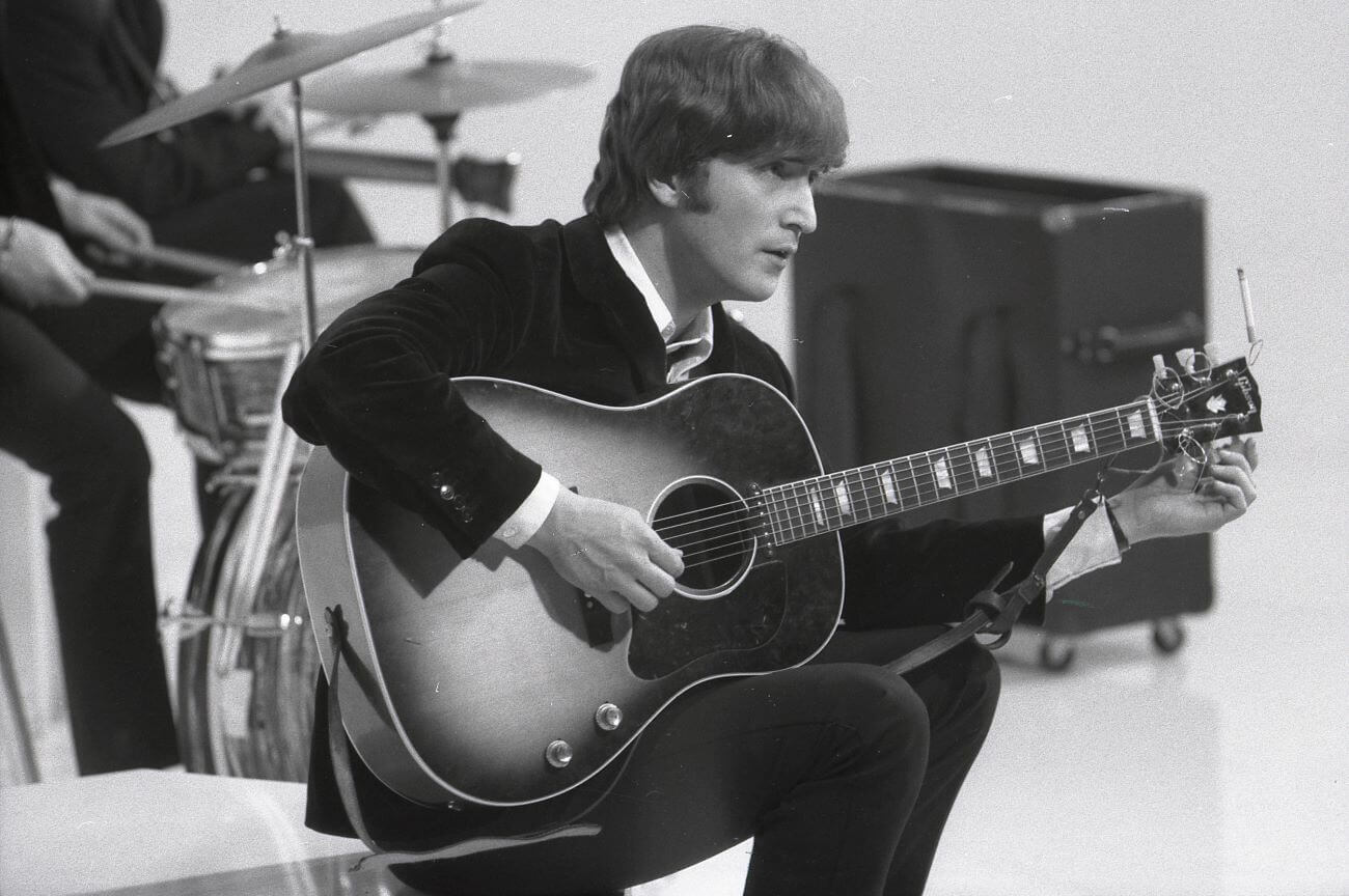 A black and white picture of John Lennon sitting on a ledge with an acoustic guitar.