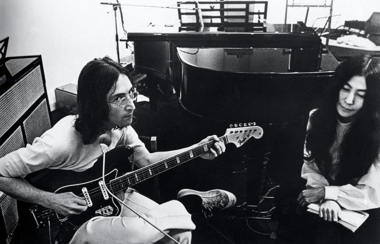Beatles member John Lennon (left) sitting and playing guitar in the studio as Yoko Ono sits next to him.