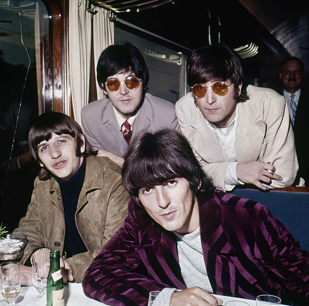 Ringo Starr and George Harrison sit at a table while Paul McCartney and John Lennon lean over the back of their seats.