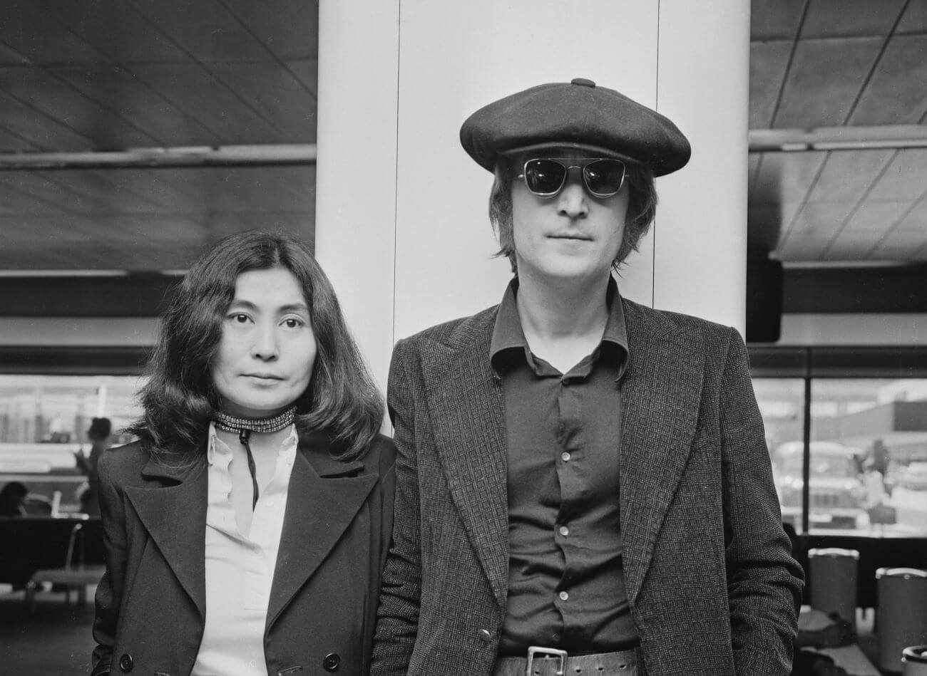 Yoko Ono and John Lennon stand next to each other at the airport. Lennon wears a hat and sunglasses.