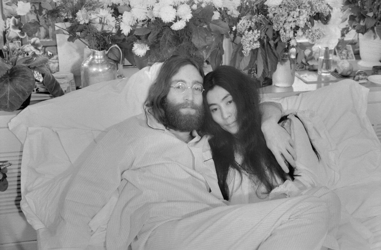 A black and white picture of John Lennon with his arm around Yoko Ono in bed wearing pajamas.