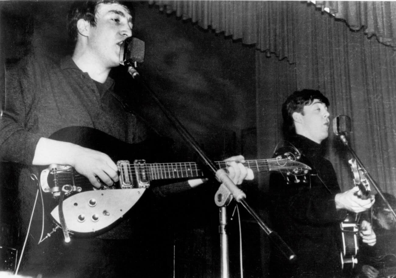 A black and white picture of John Lennon and Paul McCartney playing guitars and singing into microphones.