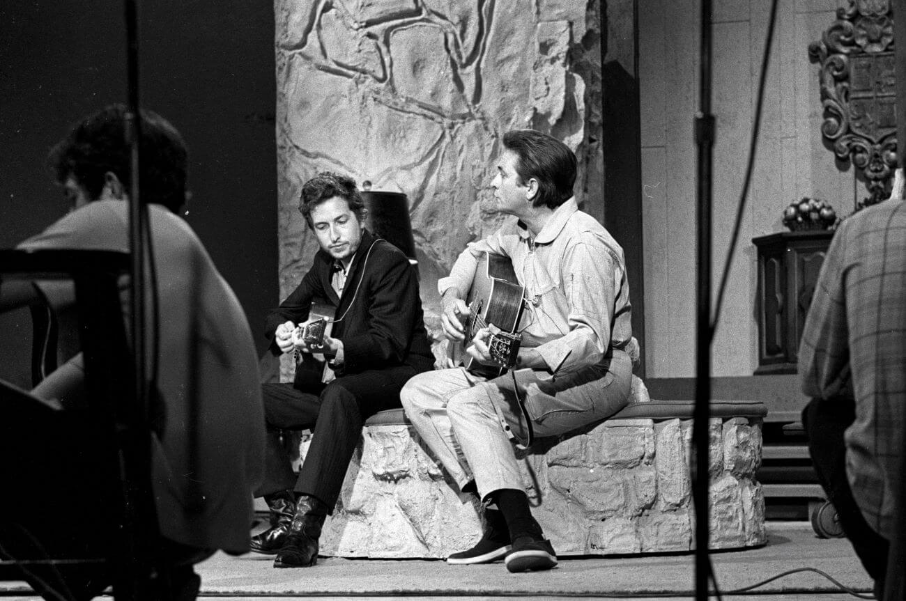 A black and white picture of Johnny Cash and Bob Dylan sitting on a ledge and playing guitars.
