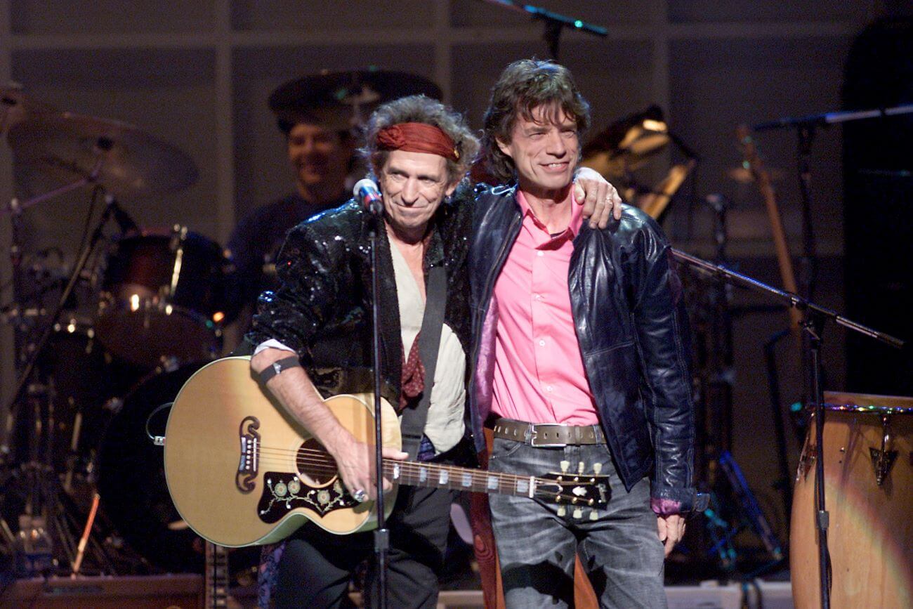 Keith Richards and Mick Jagger stand with their arms in front of each other in front of a microphone. Richards holds an acoustic guitar.