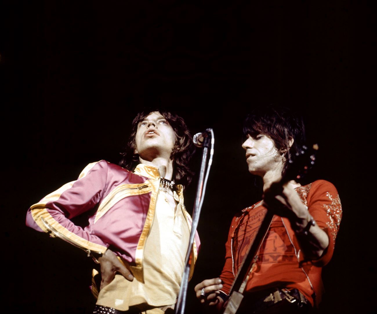 Mick Jagger stands behind a microphone with his hand on his hip. Keith Richards stands next to him, playing guitar.