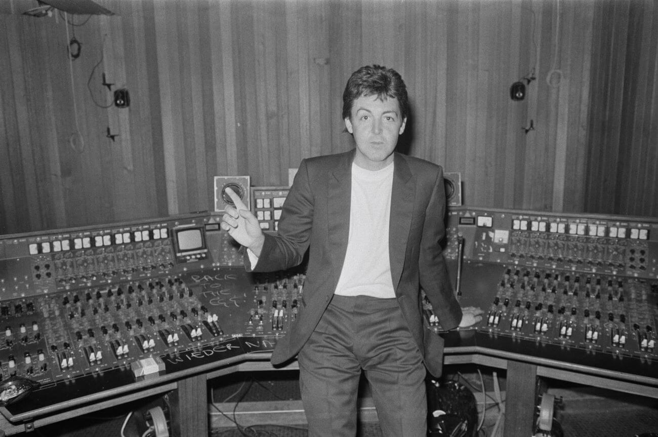 A black and white picture of Paul McCartney leaning against a recording studio control panel and holding up a hand.