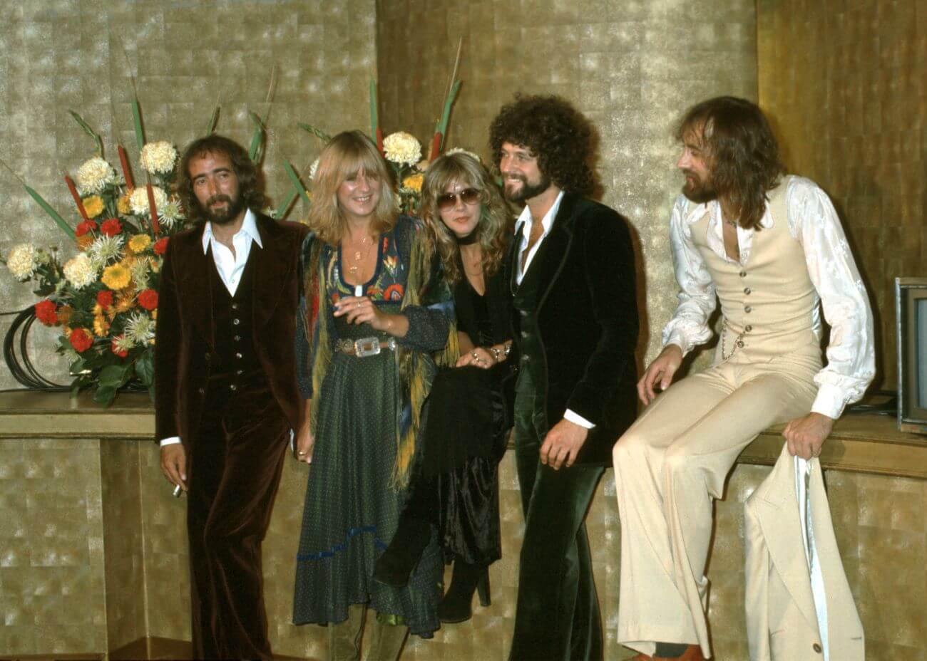 John McVie, Christine McVie, Stevie Nicks, Lindsey Buckingham, and Mick Fleetwood lean against a ledge decorated with flowers.