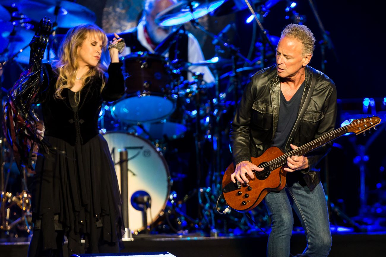 Stevie Nicks wears a black dress and dances with a tambourine. Lindsey Buckingham plays guitar across a stage from her.