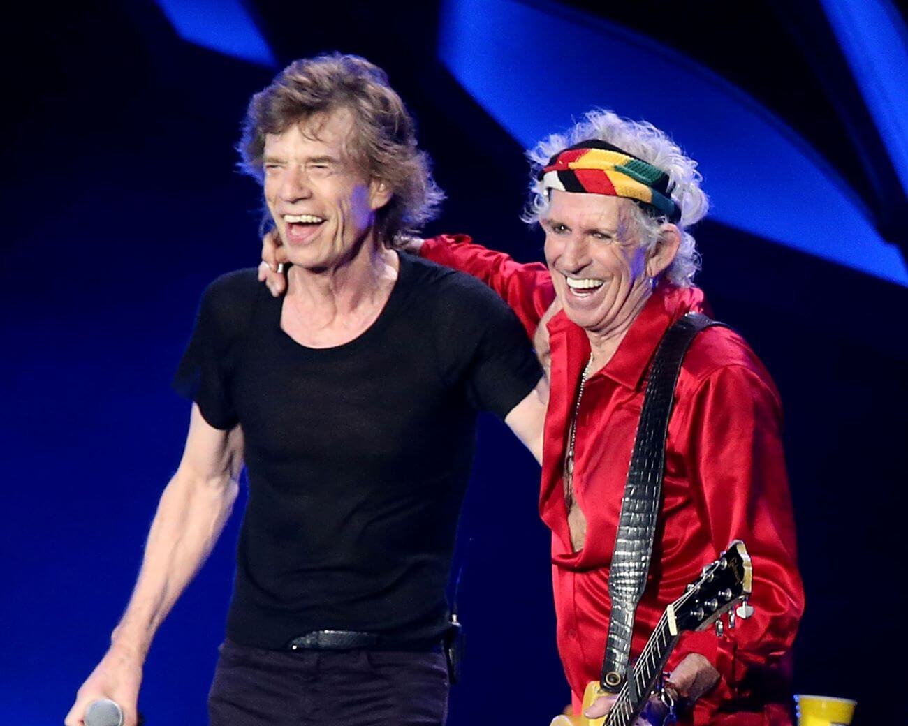 Keith Richards stands with his arm around Mick Jagger's shoulders. Jagger holds a microphone and Richards holds a guitar. 