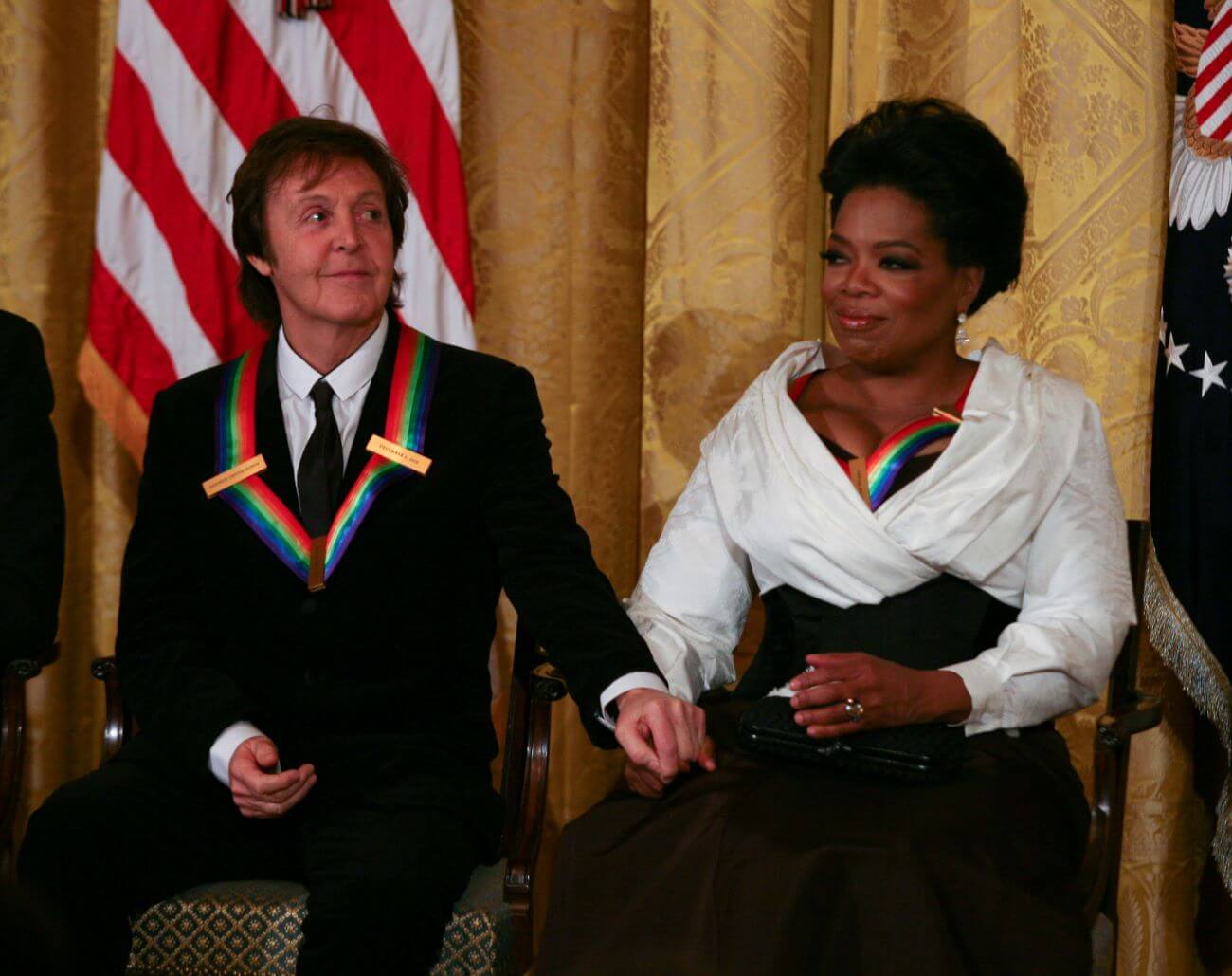 Oprah Winfrey and Paul McCartney hold hands while sitting next to each other. Both wear rainbow ribbons.
