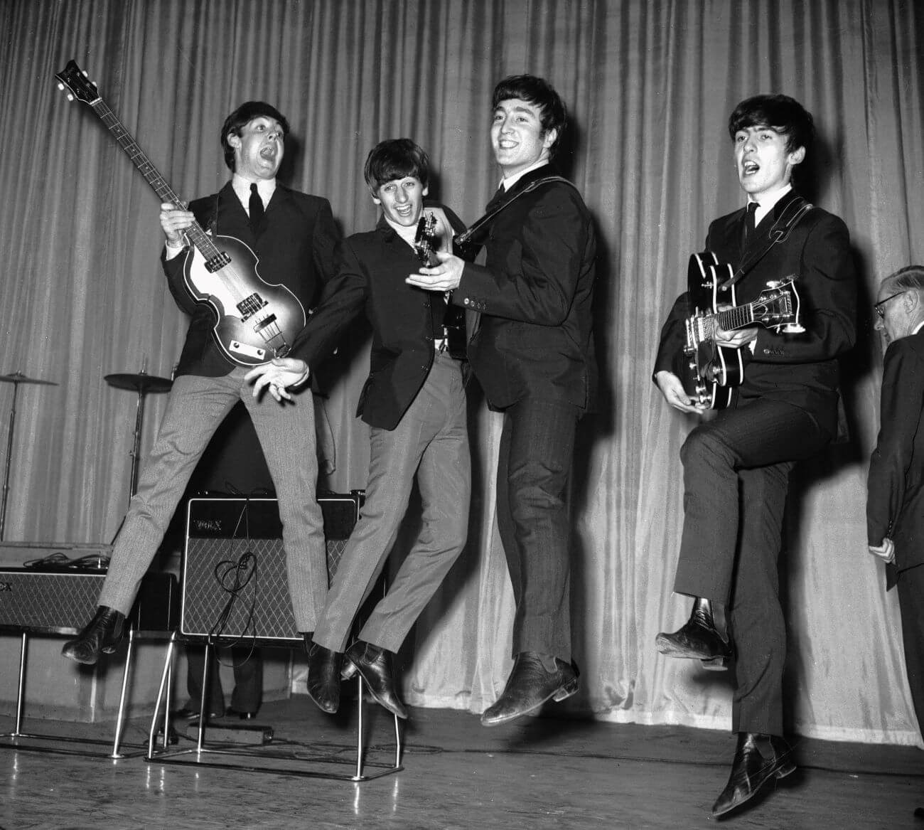 A black and white picture of Paul McCartney, Ringo Starr, John Lennon, and George Harrison jumping up in their air while on a stage. McCartney, Lennon, and  Harrison hold guitars.