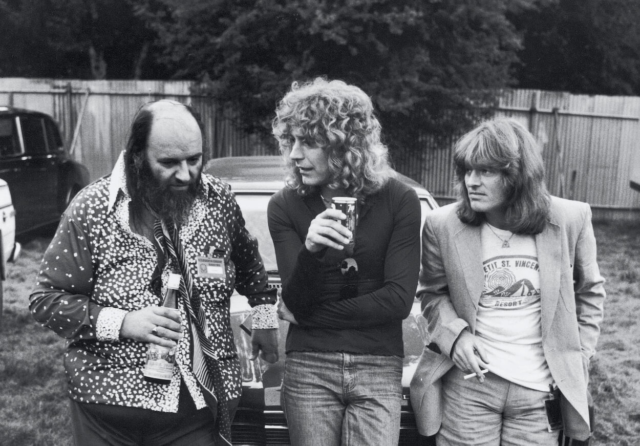 Led Zeppelin manager Peter Grant, singer Robert Plant, and bassist John Paul Jones standing and talking in front of a car in 1979.