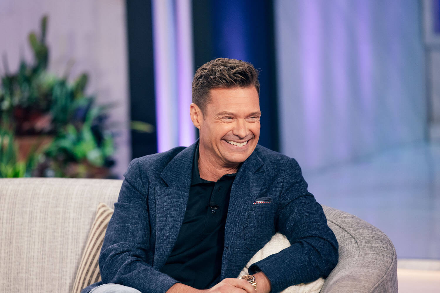 'American Idol' host Ryan Seacrest smiling on a couch. Seacrest is the future host of 'Wheel of Fortune.'