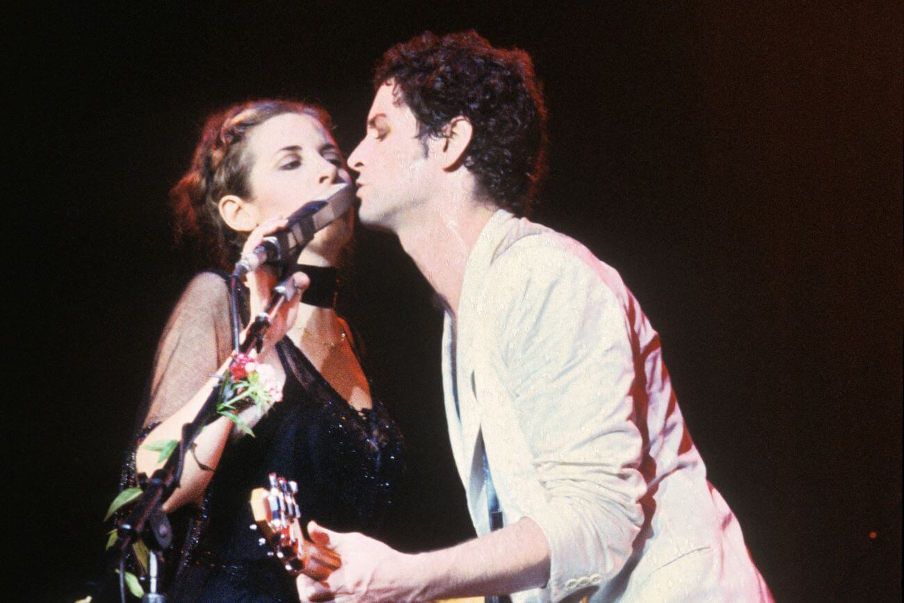 Stevie Nicks and Lindsey Buckingham sing into the same microphone while Buckingham plays guitar.
