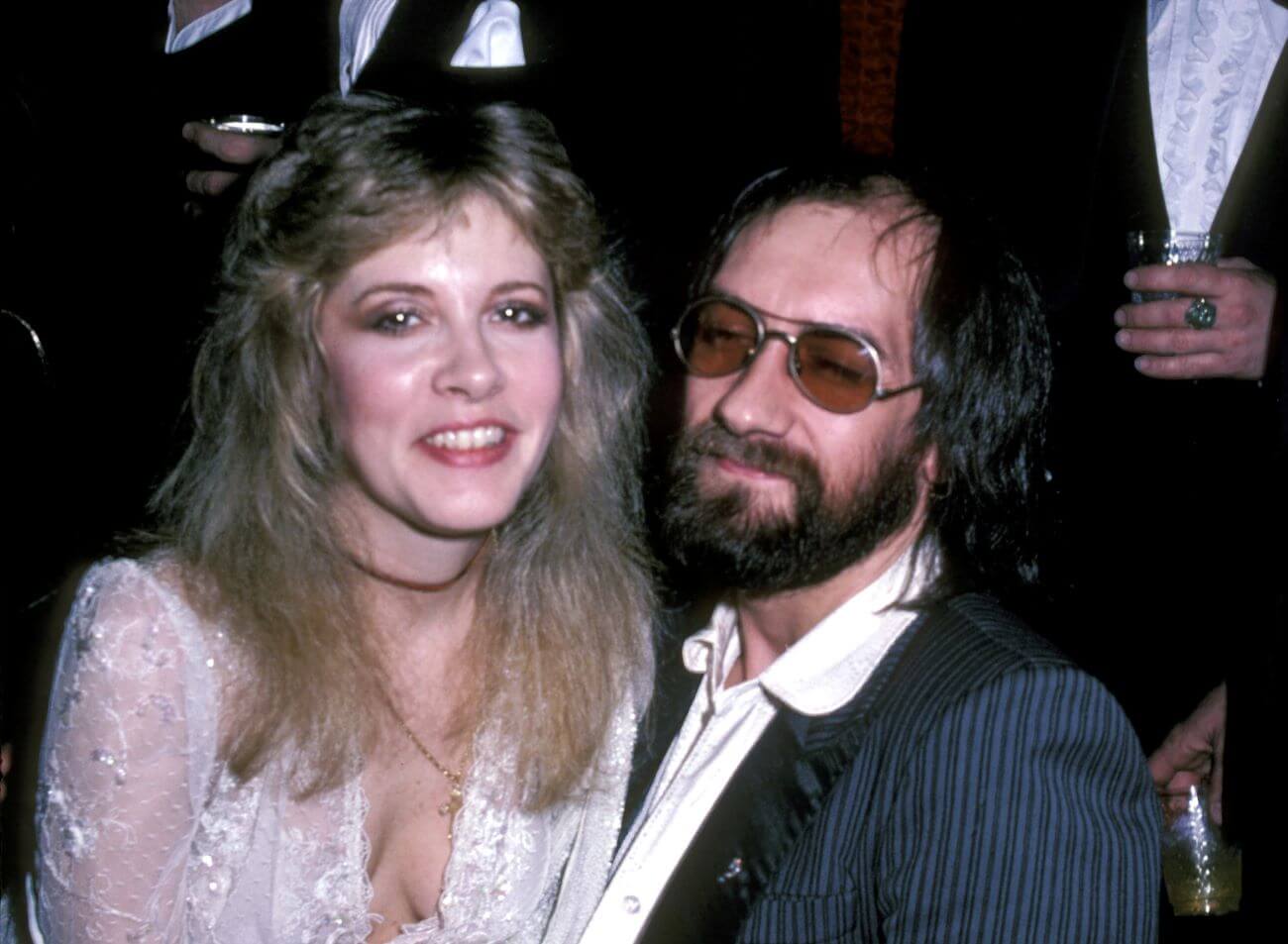 Stevie Nicks wears a white dress and sits on Mick Fleetwood's lap. He wears sunglasses.