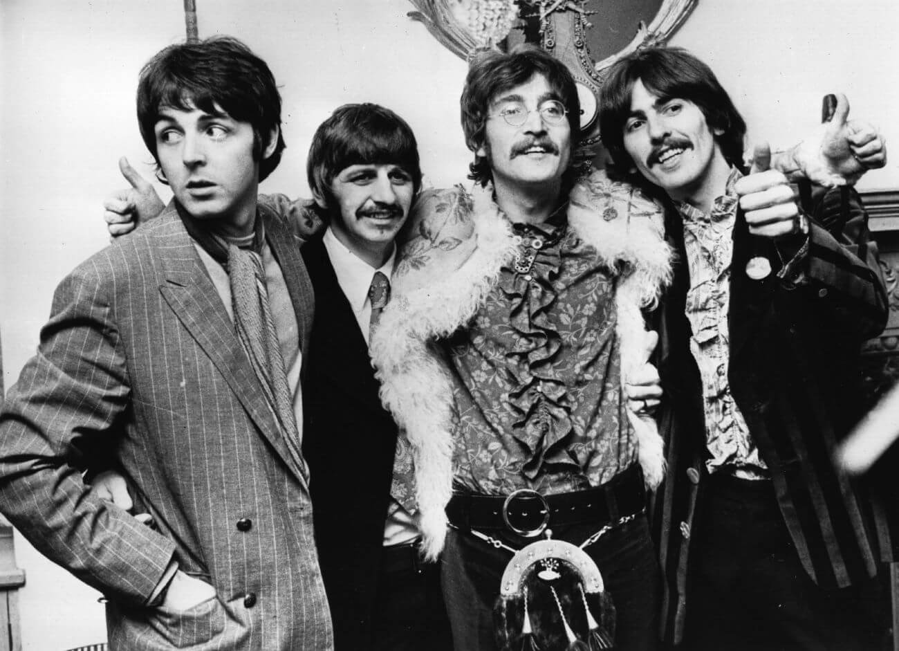 A black and white picture of Paul McCartney, Ringo Starr, John Lennon, and George Harrison posing in front of a fireplace with their arms around each other.