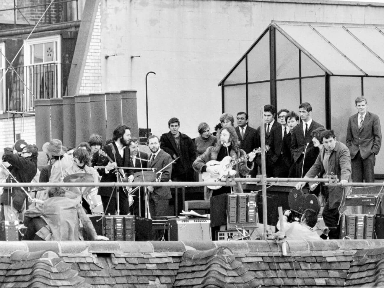 A black and white picture of The Beatles performing on a rooftop while a crew films them. 