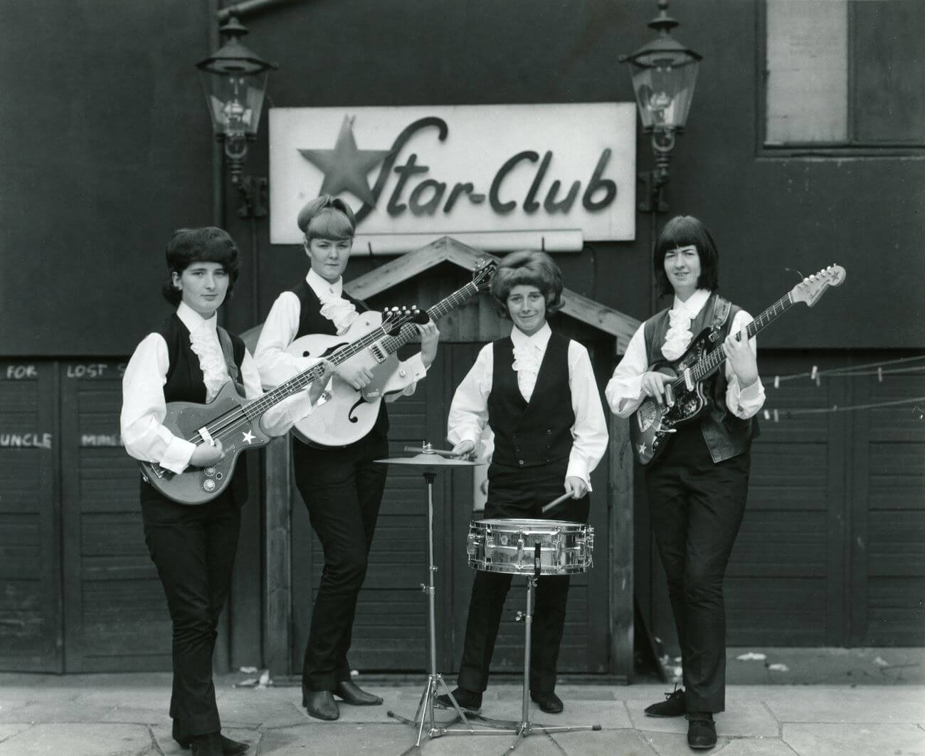 A black and white picture of Mary McGlory, Pamela Birch, Sylvia Saunders, and Valerie Gell of The Liverbirds posing with instruments outside the Star Club.