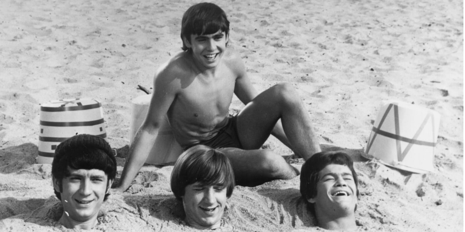 Davy Jones is seated on the sand in front of fellow 'The Monkees' co-stars Mike Nesmith, Peter Tork, and Micky Dolenz in 'Royal Flush.'