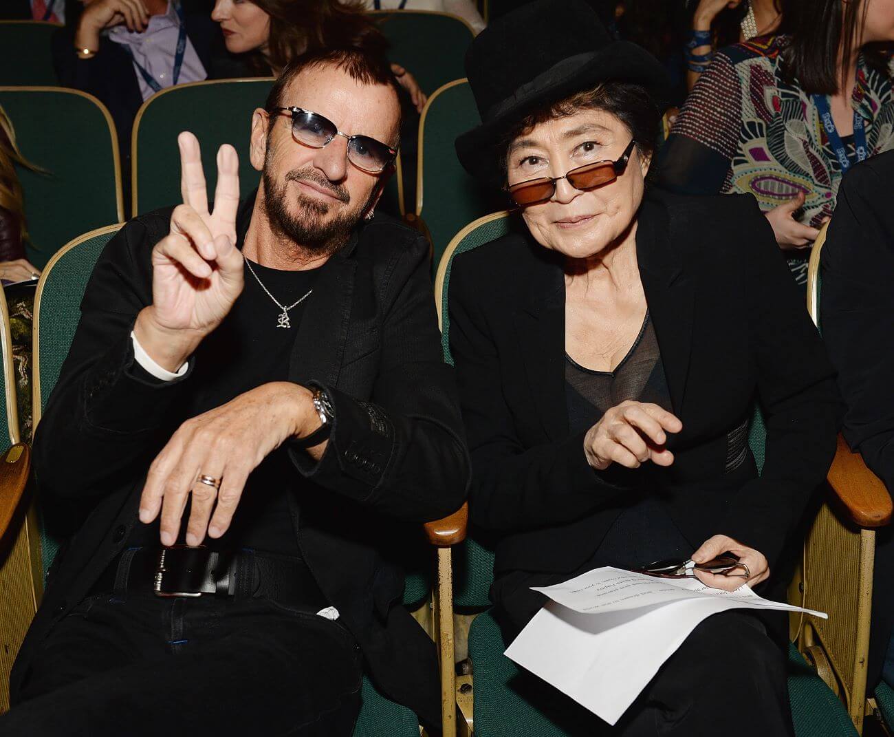 Ringo Starr wears sunglasses and holds up a peace sign. He sits next to Yoko Ono, who wears sunglasses and a black top hat.