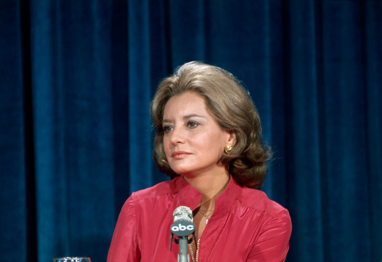 Barbara Walters wears a red shirt and sits in front of an ABC microphone. There is a blue curtain behind her.