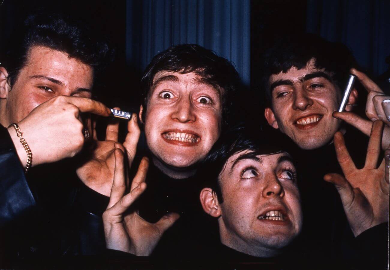 Pete Best, John Lennon, Paul McCartney, and George Harrison smile widely at the camera. McCartney holds his hands up near his face.