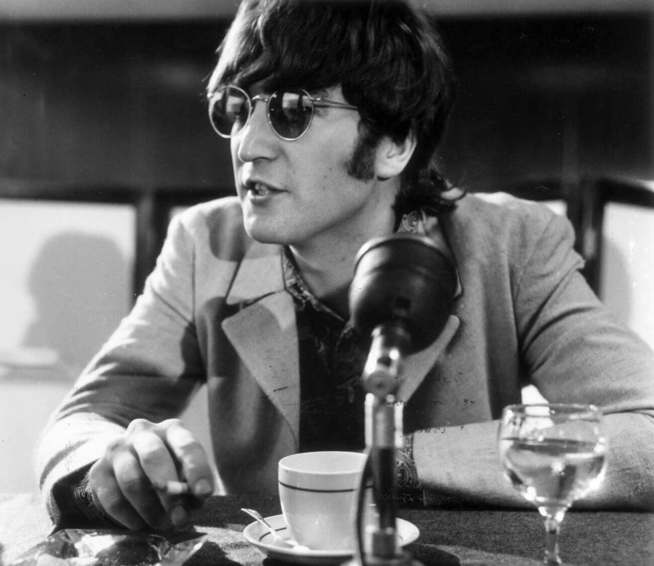 A black and white picture of John Lennon wearing sunglasses and sitting in front of a microphone, coffee cup, and water glass.