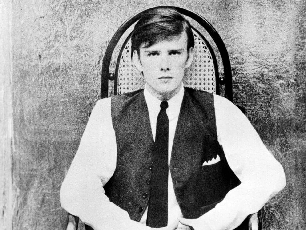 A black and white picture of The Beatles' Stuart Sutcliffe wearing a vest and tie and sitting in a chair.