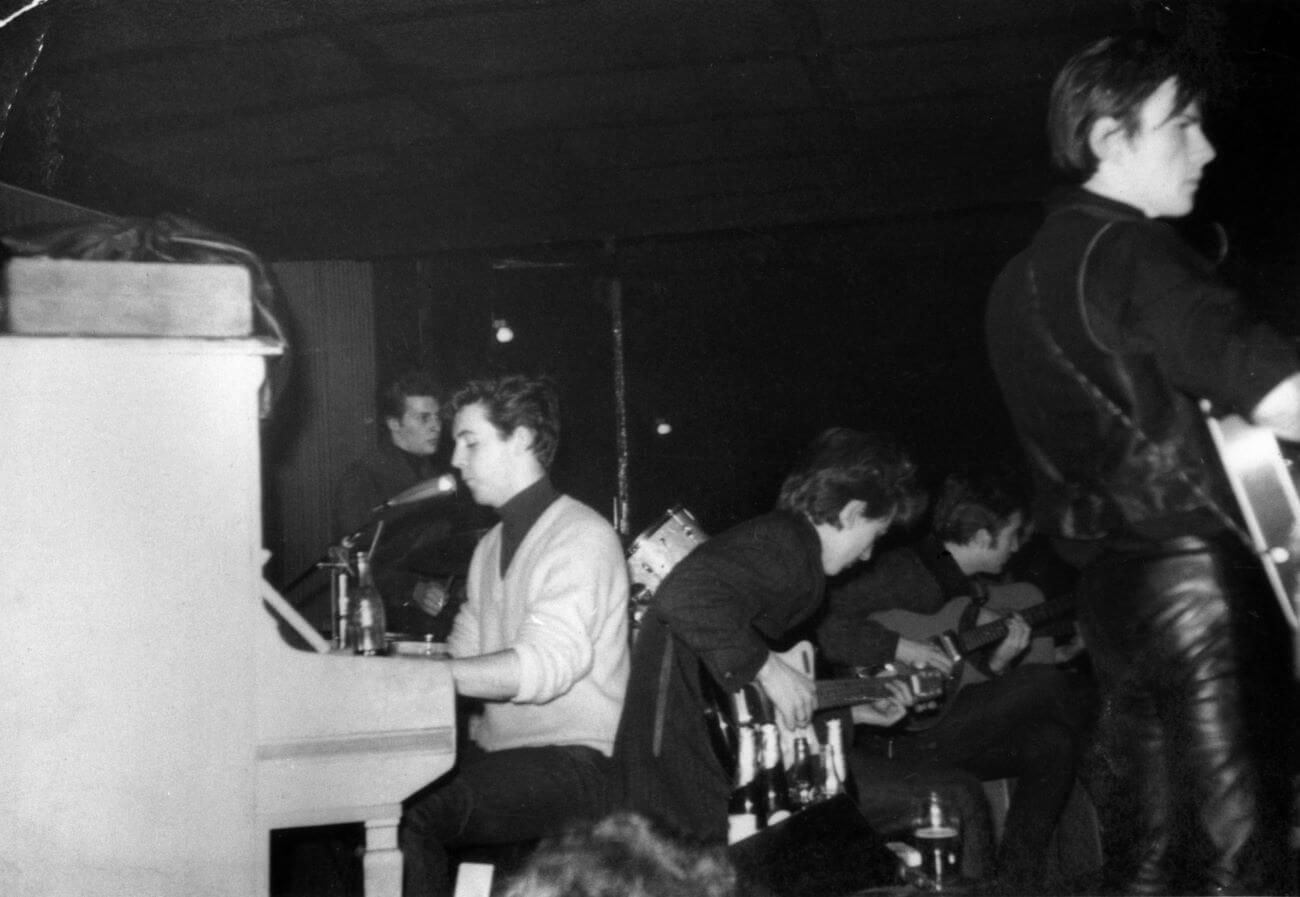 A black and white picture of The Beatles performing in Hamburg. Paul McCartney sits at the piano while Stuart Sutcliffe, George Harrison, and John Lennon play guitars.