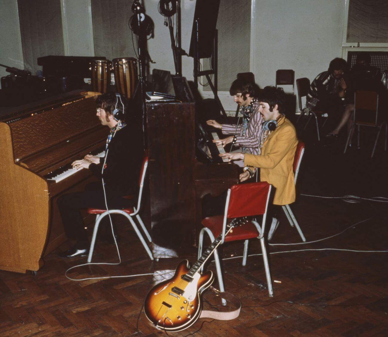 John Lennon sits at a piano wearing headphones. Paul McCartney and Ringo Starr share a piano behind Lennon.