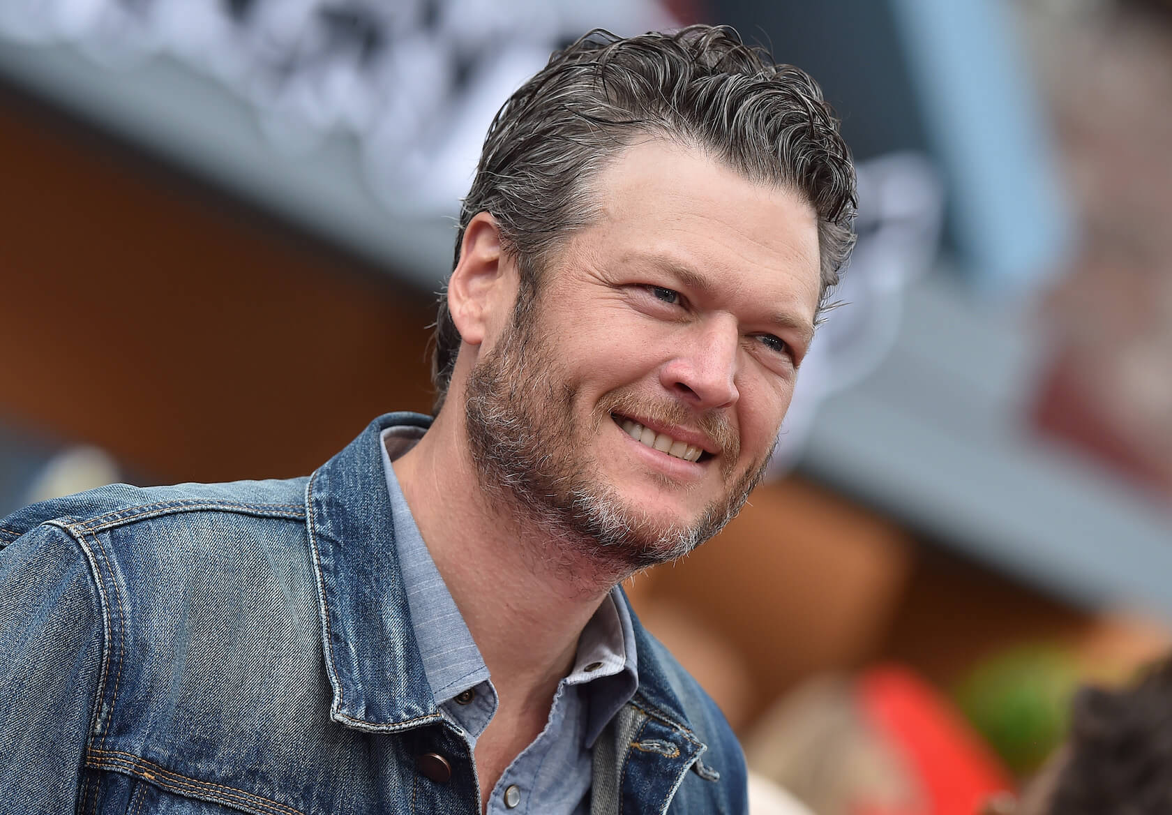 A close-up of 'The Voice' coach Blake Shelton smiling outdoors. He's wearing a denim jacket
