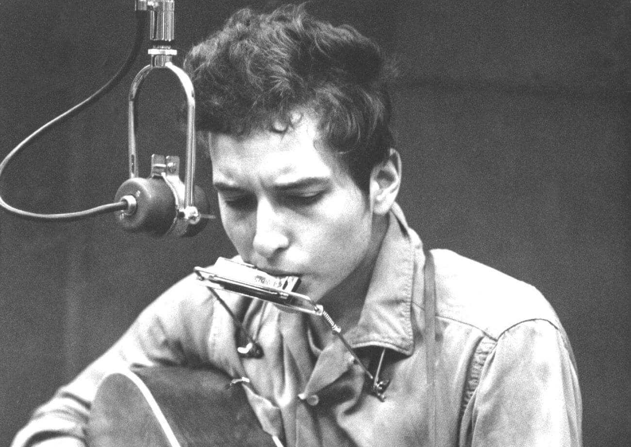 A black and white picture of Bob Dylan playing a harmonica while holding an acoustic guitar. He plays into a microphone. 