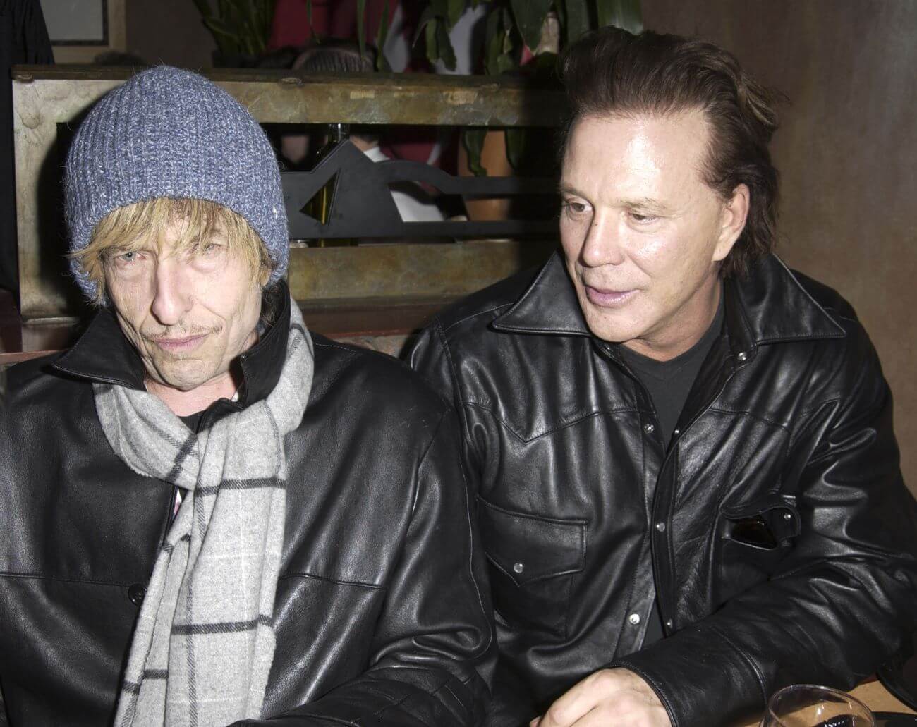 Bob Dylan and Mickey Rourke wear black leather jackets and sit together. Dylan wears a gray scarf, blue knit cap, and blonde wig.