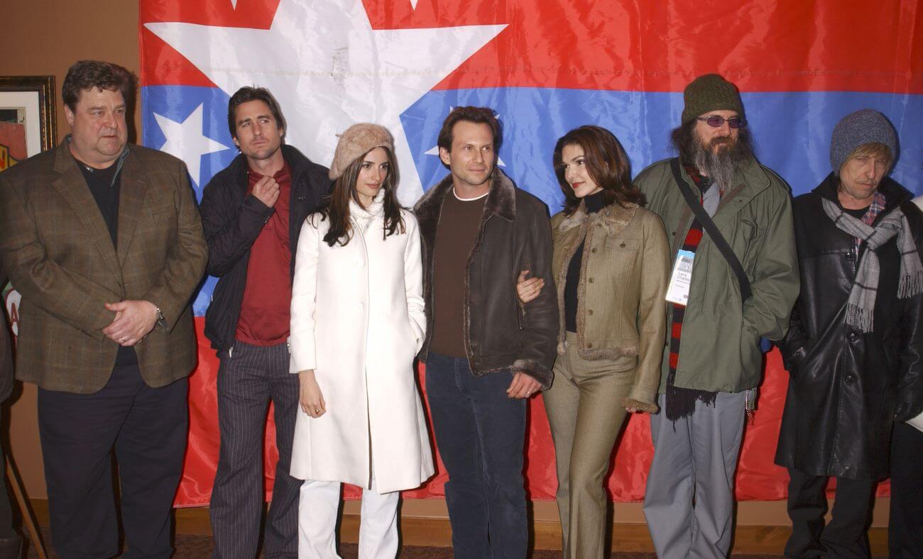 John Goodman, Luke Wilson, Penelope Cruz, Christian Slater, Laura Elena Harring, Larry Charles, and Bob Dylan wear coats and stand in front of a red and blue flag. Dylan wears a blonde wig.