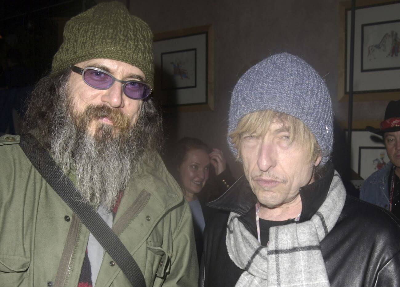 Writer Larry Charles wears sunglasses and a hat while standing with Bob Dylan, who wears a hat, blonde wig, and scarf.