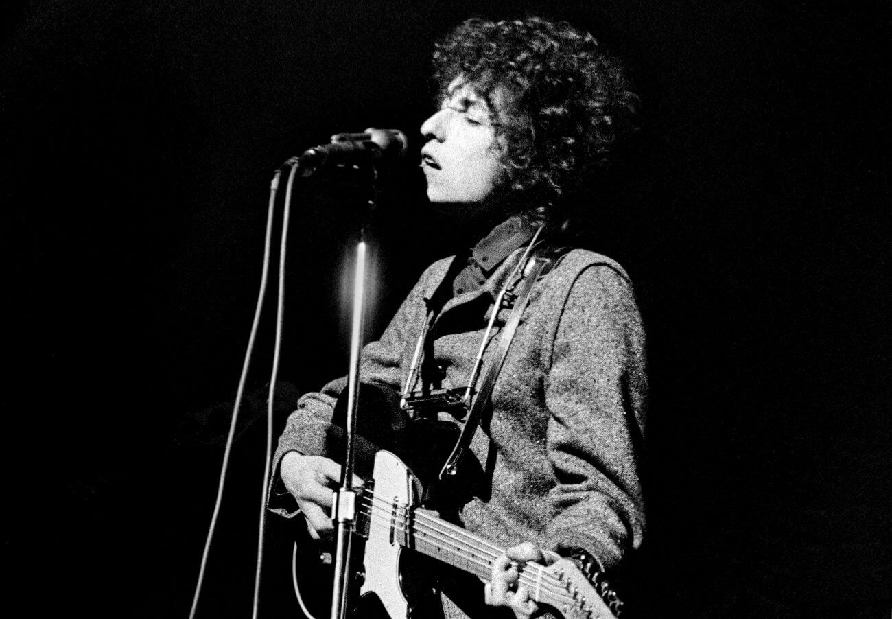A black and white picture of Bob Dylan playing an electric guitar and singing into a microphone.