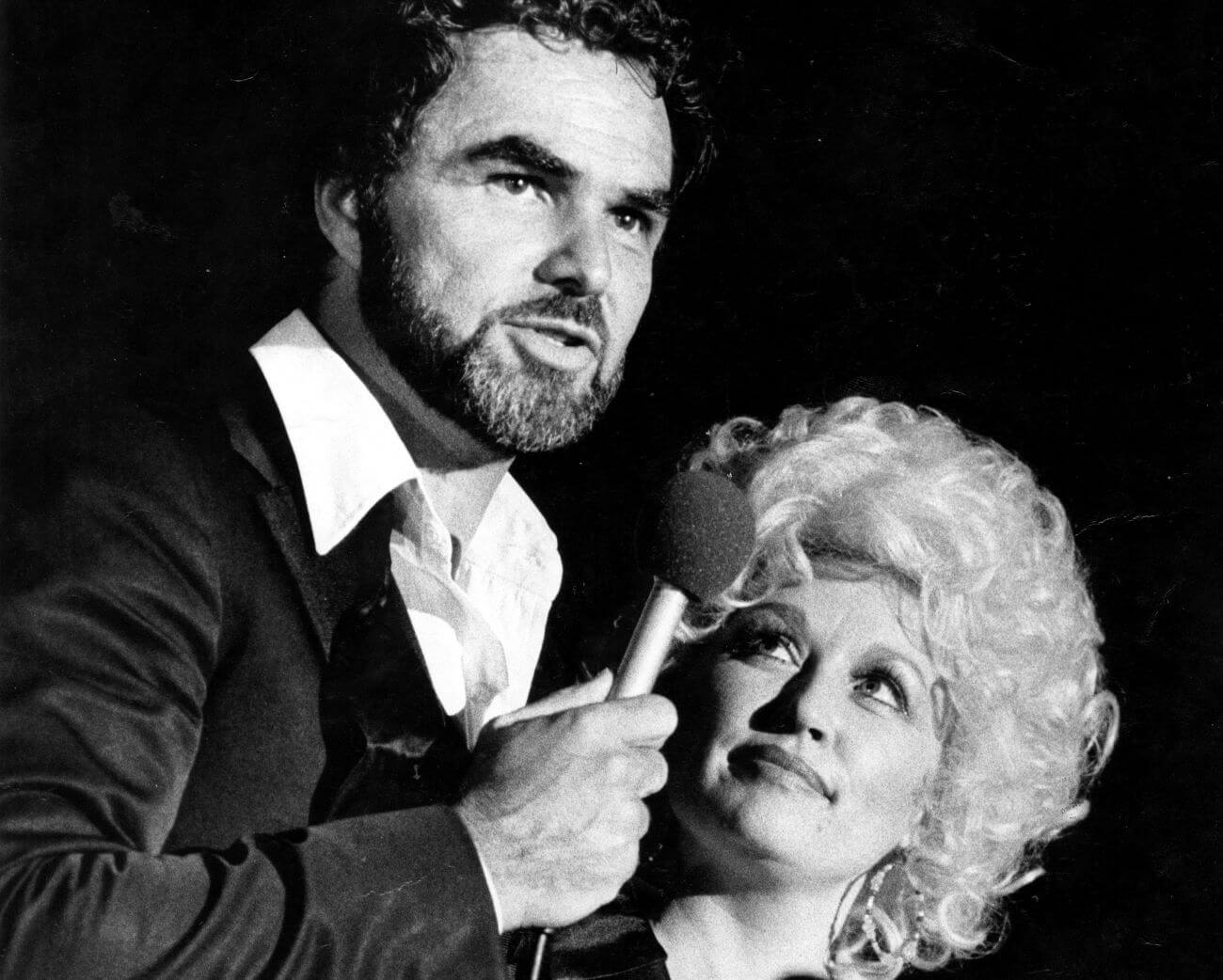 A black and white picture of Burt Reynolds speaking into a microphone while Dolly Parton looks up at him.