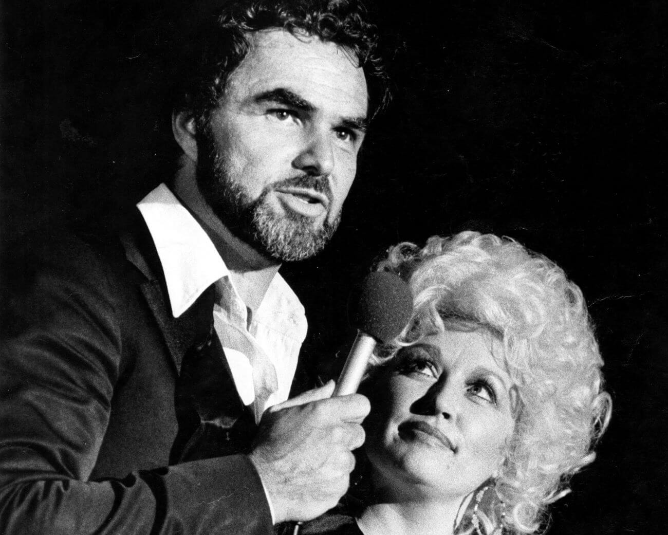 A black and white picture of Burt Reynolds speaking into a microphone while Dolly Parton looks up at him.