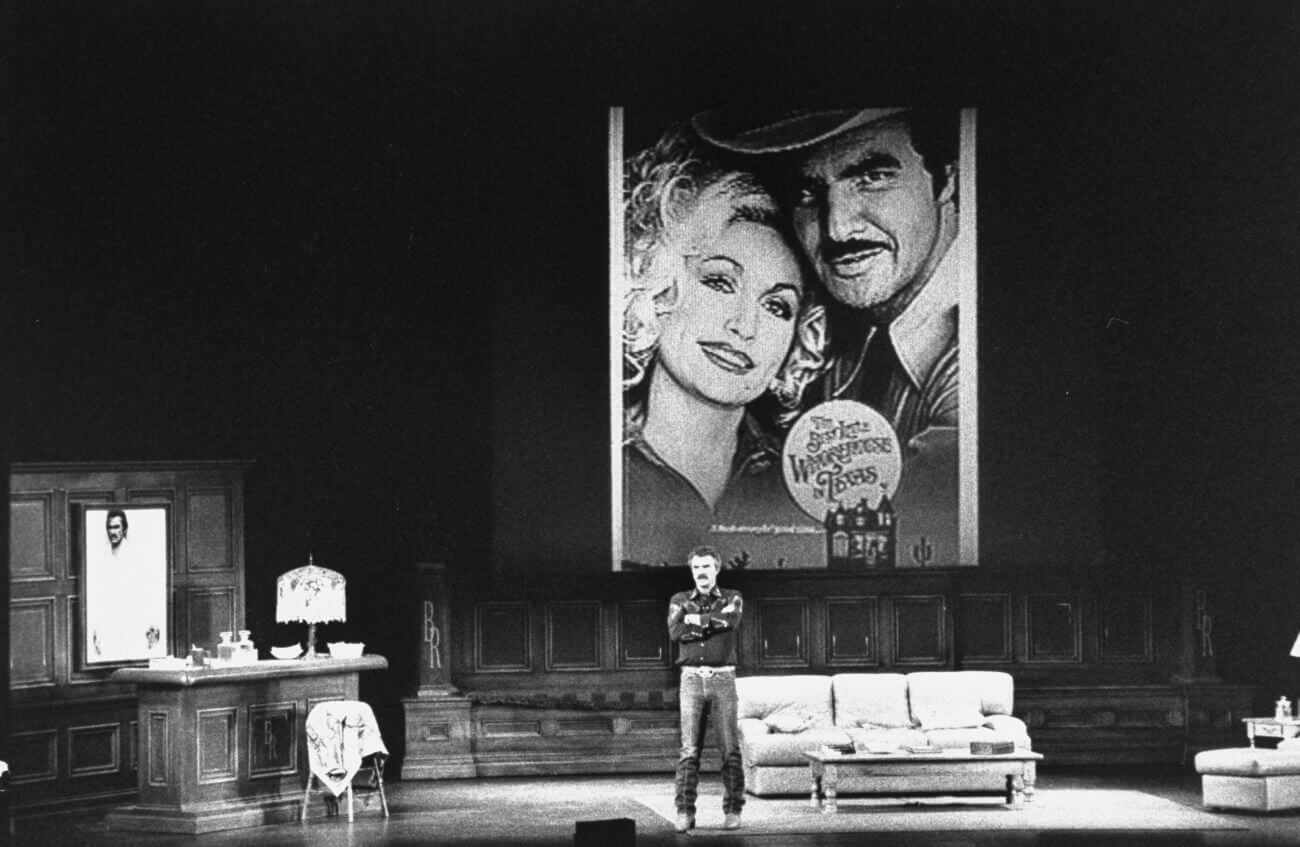 A black and white picture of Burt Reynolds standing on a stage with a couch and bar. There's a large poster of him and Dolly Parton for 'The Best Little Whorehouse in Texas' behind him.