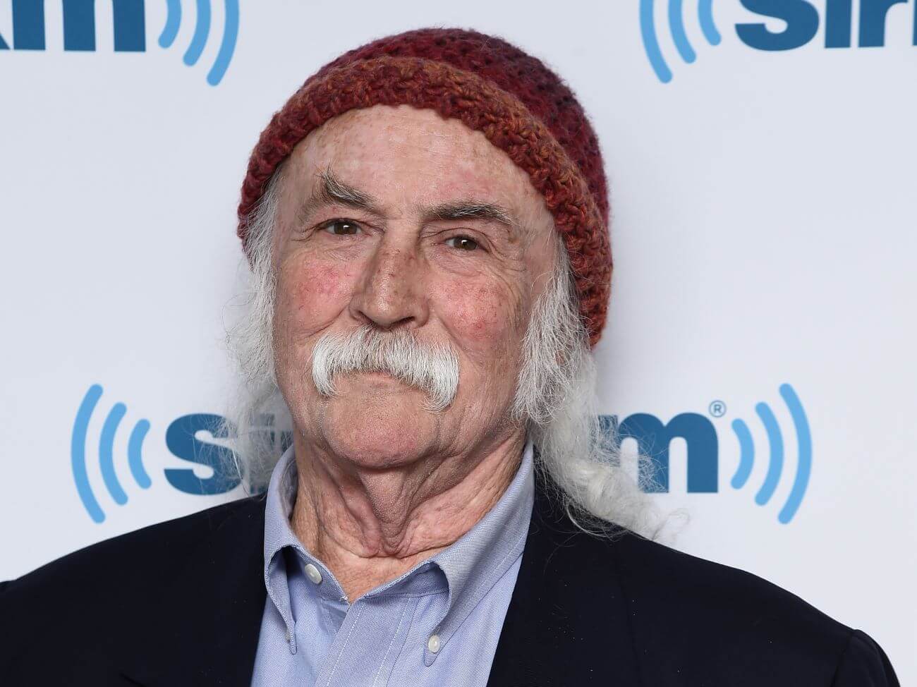 David Crosby wears a collared shirt, black jacket, and red knit hat. He stands in front of a sign for "Sirius XM."