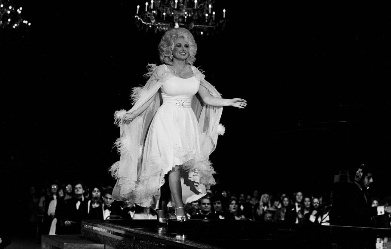 A black and white picture of Dolly Parton wearing a dress and cape and walking on a stage.