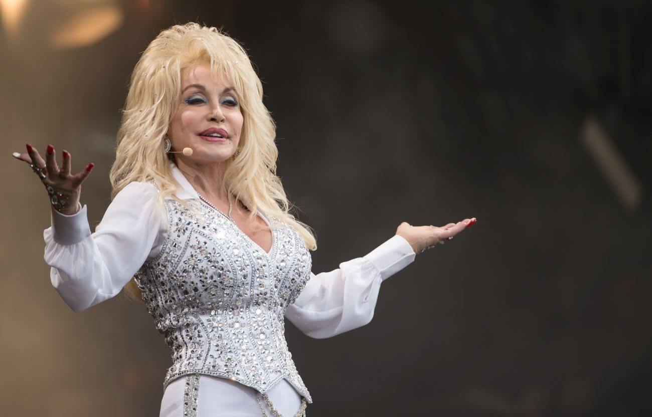 Dolly Parton wears a white sequined shirt and matching pants. She lifts her arms in the air.