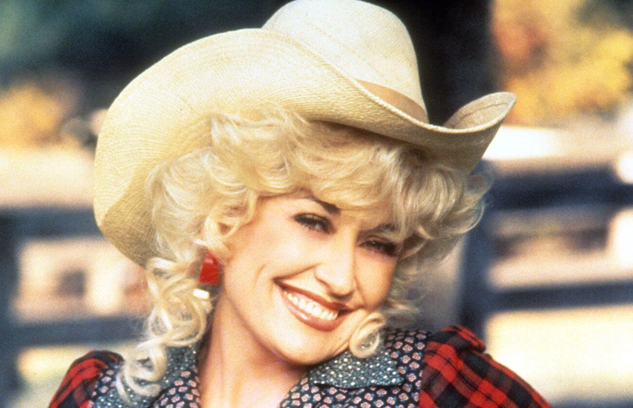 Dolly Parton wears a cowboy hat and stands in front of a fence.