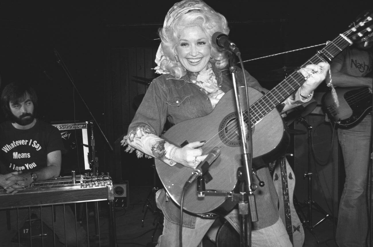A black and white picture of Dolly Parton wearing a denim shirt and head scarf while playing an acoustic guitar. She stands in front of a microphone.