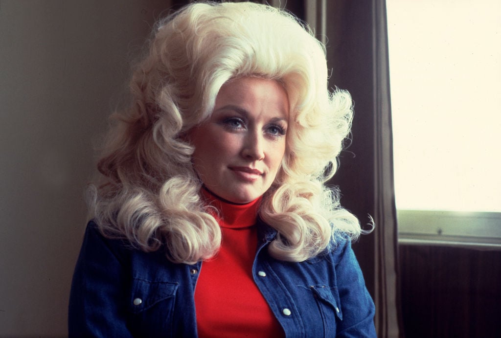 Dolly Parton sitting down in a red turtleneck and jean shirt.