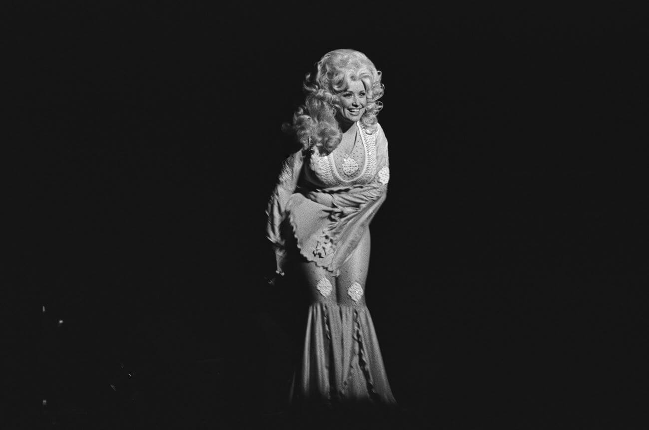 A black and white picture of Dolly Parton wearing a jumpsuit and leaning forward. She holds her arm across her body.