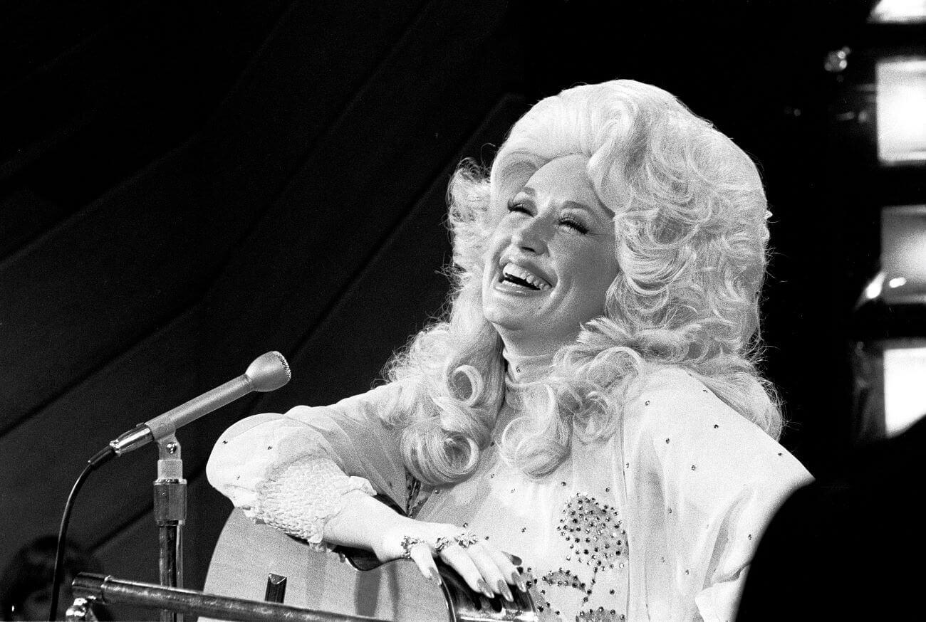 A black and white picture of Dolly Parton laughing and resting an acoustic guitar on her lap. She sits in front of a microphone.