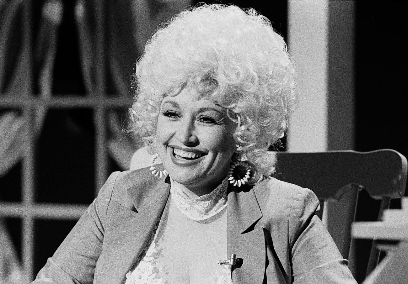 A black and white picture of Dolly Parton sitting in a rocking chair in front of a window.